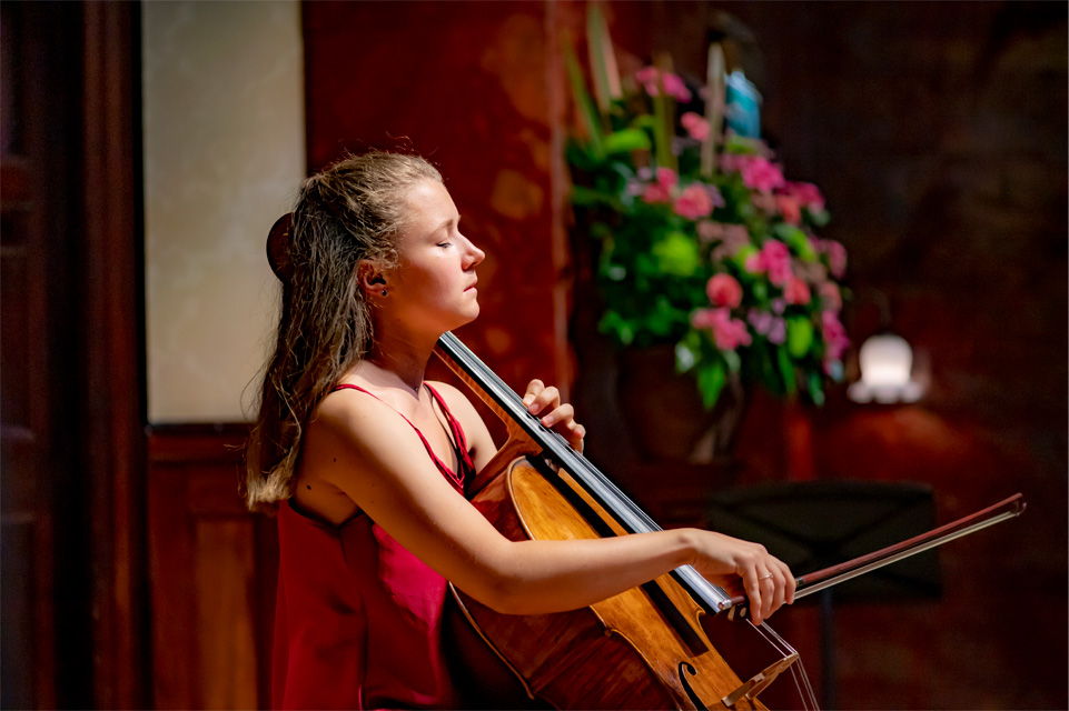 A female student, wearing a red dress, playing the cello solo on stage.